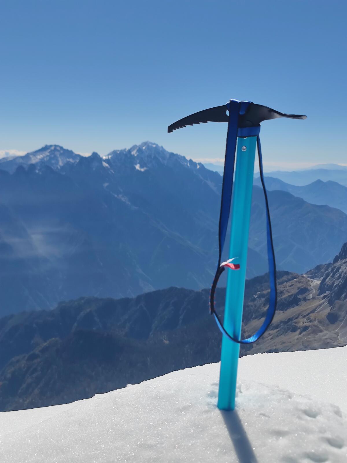 An ice axe planted in the summit snow of Haba Snow Mountain, with Jade Dragon Snow Mountain visible on the horizon under a clear blue sky.