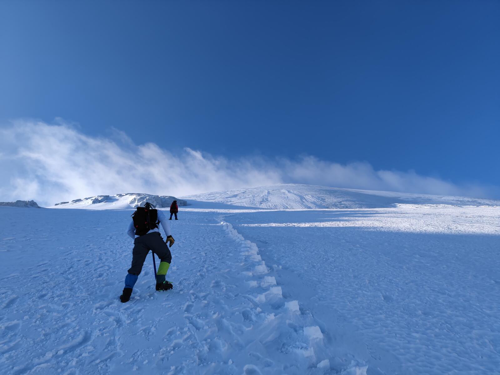 A solo climber following a bootpack track across the exposed final approach near the summit, wind blowing snow off the ridge under a deep blue sky.